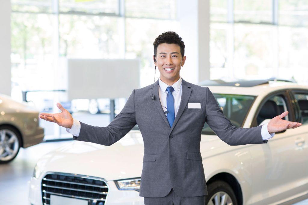 Car salesman standing with his arms stretched out in front of a car